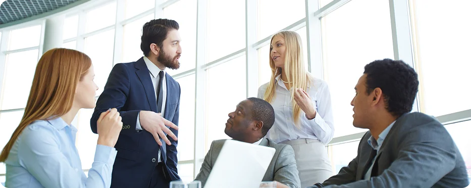 A group of five professionals engaged in a discussion during a meeting in a bright office space with large windows.