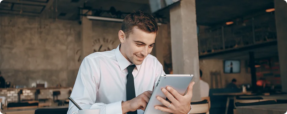 A man in a white shirt and tie sits at a café table, focused on a tablet in his hands, with a modern industrial background.