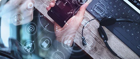 A person holding a smartphone, with a keyboard and headset beside them, surrounded by digital communication icons.