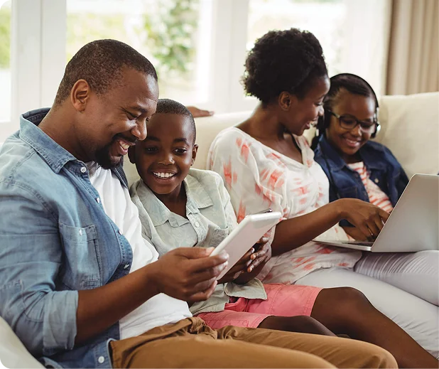 A family sits together on a couch, engaged with various devices, including a tablet and a laptop, in a bright living room.