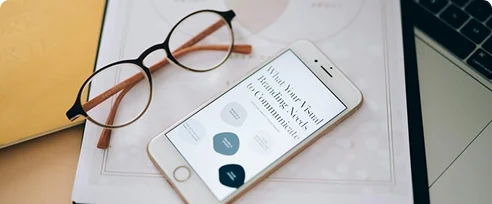 A smartphone displaying a text on visual communication, alongside a pair of glasses and a laptop, on a desk with a notebook.