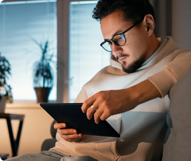 A person sits on a couch, attentively holding a tablet, to check the Benefits of Business VoIP..